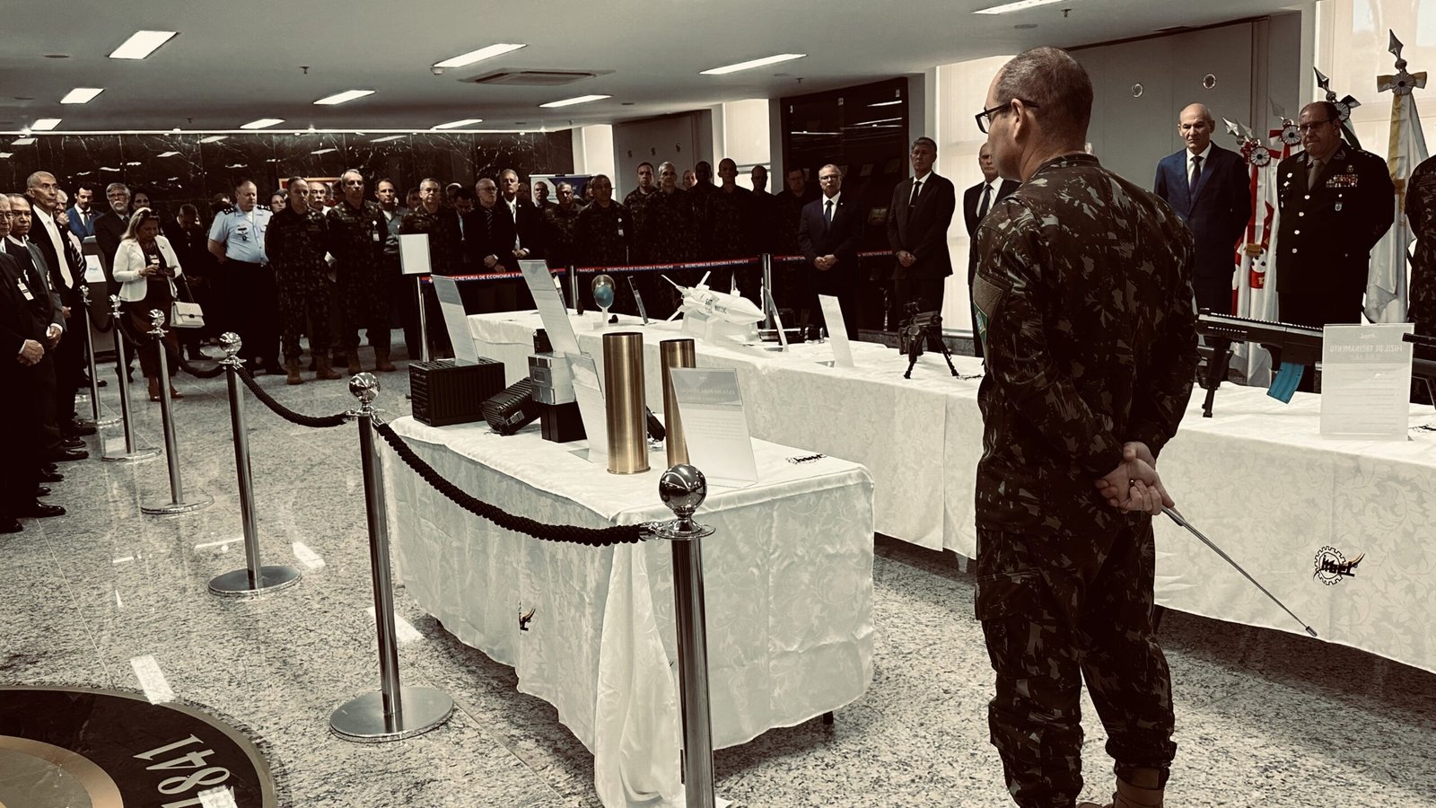 Visão geral da exposição IMBEL no Espaço Gen. Leônidas Pires Gonçalves — Brasília, 1º de julho de 2026. Ao fundo, generais e autoridades civis assistem à apresentação dos produtos. Crédito: Acervo Reaction News.